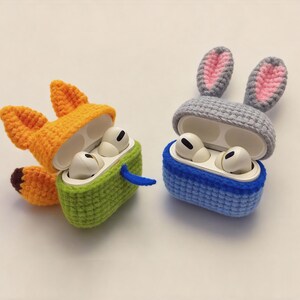 Two crochet AirPod cases: one shaped like a fox with orange and green accents, and the other a blue and grey bunny. The fox case has orange ears and a brown tail. The bunny case has pink inner ears.