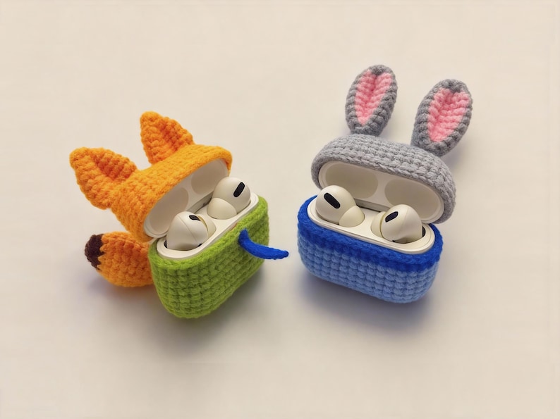 Two crochet AirPod cases: one shaped like a fox with orange and green accents, and the other a blue and grey bunny. The fox case has orange ears and a brown tail. The bunny case has pink inner ears.