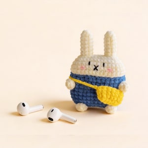 A crochet bunny-shaped case in white, blue, and yellow, designed to hold a pair of white wireless earbuds. The bunny has a blue outfit and a yellow bag. The earbuds are lying on a light yellow surface.