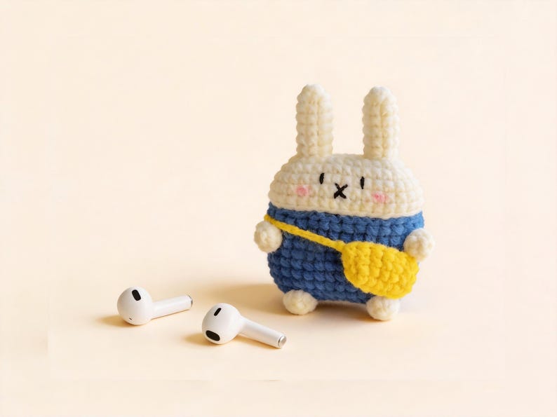 A crochet bunny-shaped case in white, blue, and yellow, designed to hold a pair of white wireless earbuds. The bunny has a blue outfit and a yellow bag. The earbuds are lying on a light yellow surface.