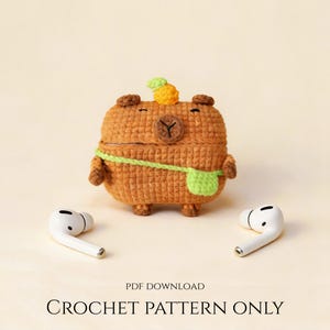 Peut inclure: Un étui marron crocheté en forme de capybara, avec une sangle verte et une petite ananas sur la tête. Deux écouteurs sans fil blancs sont de chaque côté de l'étui. Le texte "PDF DOWNLOAD CROCHET PATTERN ONLY" est en bas.