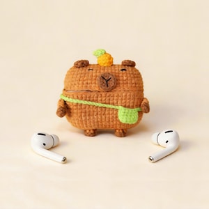 A crocheted brown capybara-shaped case for wireless earbuds, with a green strap and a small pineapple on its head. Two white earbuds are on either side of the case. The case is a unique accessory.