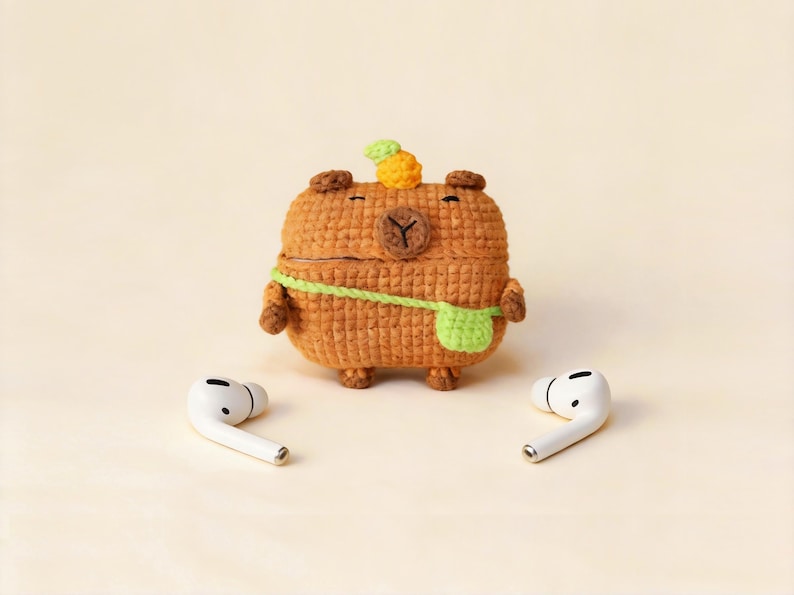 A crocheted brown capybara-shaped case for wireless earbuds, with a green strap and a small pineapple on its head. Two white earbuds are on either side of the case. The case is a unique accessory.