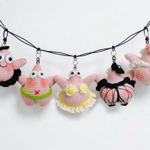 Puede incluir: Una guirnalda con cinco figuras de estrellas de mar tejidas a ganchillo en color rosa, suspendidas de un cordón negro. Cada figura luce un atuendo distinto, incluyendo un cuello de volantes negro, un cinturón de sumo verde y un tutú blanco.