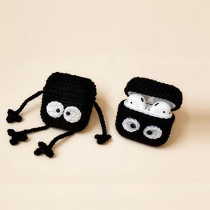 Two black crochet AirPod cases with white eyes and arms and legs. One case is open, revealing white Airpods. The other case is lying down. The cases are on a light beige background.