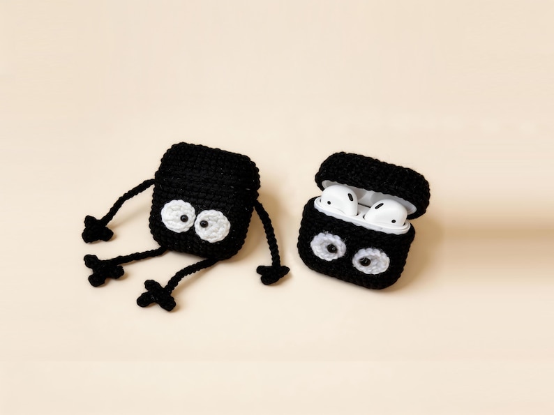 Two black crochet AirPod cases with white eyes and arms and legs. One case is open, revealing white Airpods. The other case is lying down. The cases are on a light beige background.