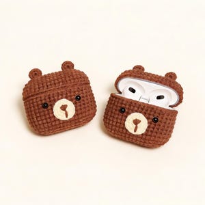 Two brown crochet bear-shaped AirPod cases. One case is closed, and the other is open, revealing white AirPods. Each case has black eyes and a cream-coloured nose. The cases have small ear details.