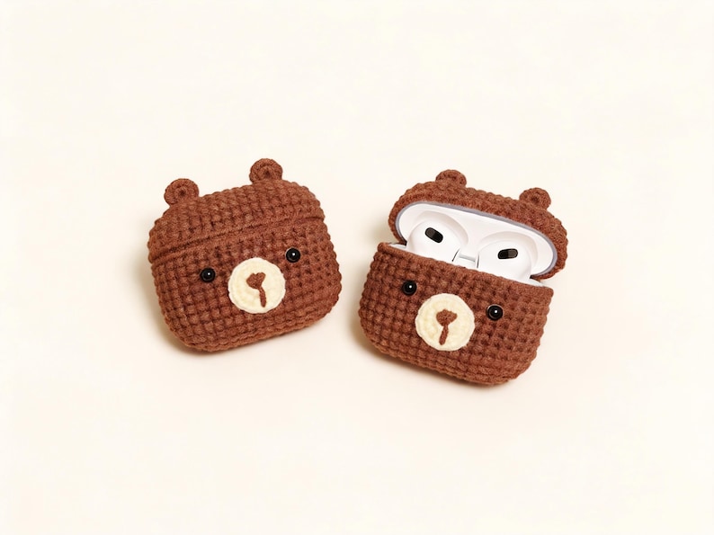 Two brown crochet bear-shaped AirPod cases. One case is closed, and the other is open, revealing white AirPods. Each case has black eyes and a cream-coloured nose. The cases have small ear details.