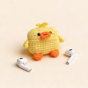 A yellow crocheted duck-shaped case for wireless earbuds, with orange feet and beak, and black eyes. Two white wireless earbuds are on either side of the case. The case has small yellow wings and a small yellow crest.