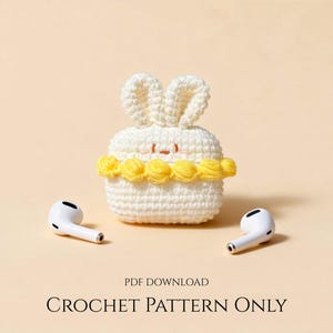 May include: A white crochet bunny-shaped case with yellow accents, designed to hold earbuds. The case has a smiling face and bunny ears. Two white earbuds are placed on either side of the case. The text "CROCHET PATTERN ONLY" is at the bottom.
