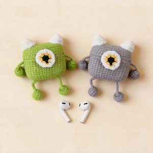 Two crochet monster-shaped AirPod cases, one green and one grey, each with a white and yellow eye design. The cases have white horns, arms, and legs. Two white AirPods are placed below the cases.