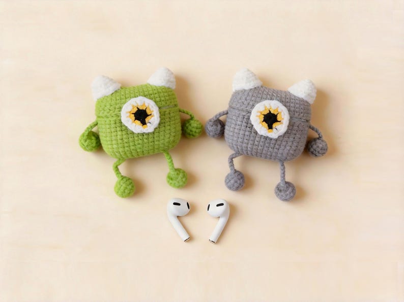 Two crochet monster-shaped AirPod cases, one green and one grey, each with a white and yellow eye design. The cases have white horns, arms, and legs. Two white AirPods are placed below the cases.