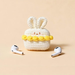 A white, crocheted bunny-shaped case for wireless earbuds, with yellow floral accents and a small face. Two white earbuds are placed on either side of the case. The background is a solid, light beige colour.