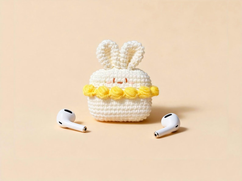 A white, crocheted bunny-shaped case for wireless earbuds, with yellow floral accents and a small face. Two white earbuds are placed on either side of the case. The background is a solid, light beige colour.