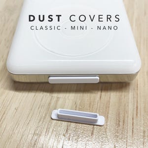 May include: A white plastic dust cover for electronic devices, featuring a rectangular shape with a small protruding tab. The text DUST COVERS CLASSIC - MINI - NANO is printed above the device, which has one cover installed and one loose.