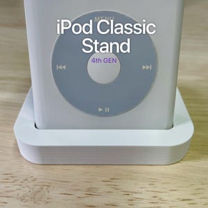 May include: A white plastic stand designed for an iPod Classic, featuring a rectangular base with a central slot. The device inserted into the stand displays a circular control interface with the text iPod Classic Stand and 4th GEN.