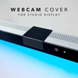 May include: A black plastic webcam cover for a Studio Display, featuring a textured top surface. The cover is positioned over the camera lens area of a silver and black monitor. Text above the device reads: WEBCAM COVER FOR STUDIO DISPLAY.