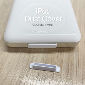 May include: A white plastic dust cover for an iPod, featuring the text iPod Dust Cover CLASSIC + MINI. The device is shown with one white rectangular port plug inserted into the bottom and a second identical white plug resting separately below it.