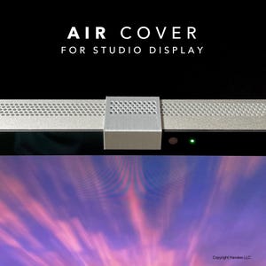 May include: A silver-coloured air cover for Studio Display featuring a perforated top surface. The text AIR COVER FOR STUDIO DISPLAY is printed above the device. A small green light is visible on the black bezel of the display screen below.