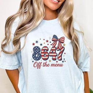 May include: A light blue t-shirt featuring a graphic with the number 8647 in a red, white, and blue American flag pattern, accented by a matching bow and stars. Below the number, the text reads Off the menu in a red, cursive script font.