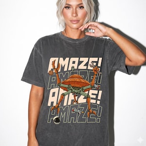 May include: A charcoal gray short-sleeve t-shirt featuring a graphic of an orange UFO with mechanical legs holding a wrench. The text AMAZE! is repeated in the background in a light cream color. Small illustrations of a planet, rocket, and helmet are included.