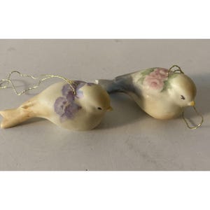 May include: Two ceramic bird ornaments. One bird has purple floral accents, the other has pink floral accents. Both birds have a cream-colored body, yellow beaks, and gold string for hanging.