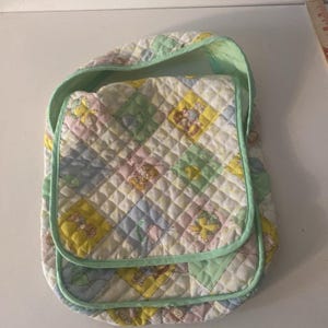 May include: A quilted baby bag with a flap closure and a shoulder strap. The bag features a patchwork design with pastel squares in yellow, pink, blue, and green, each with a cartoon character. The bag is trimmed with a light green binding.