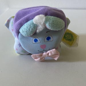 May include: A vintage sheep-shaped plush toy with a light blue face, blue eyes, and a pink bow tie. It wears a purple hat and has light blue and white accents. The toy has a tag attached.