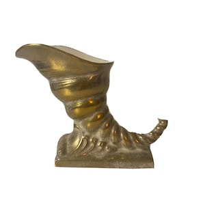 May include: A brass-coloured cornucopia-shaped decorative object. The object has a curved, spiralling body with a flared opening at the top. The base is rectangular with decorative flourishes. The overall design is ornate and vintage.