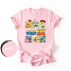 May include: A light pink short-sleeve t-shirt featuring a graphic print of Toy Story characters including Woody, Buzz Lightyear, Jessie, the Aliens, Bullseye, Hamm, Mr. Potato Head, Slinky Dog, and Rex, with the Toy Story logo in the center.