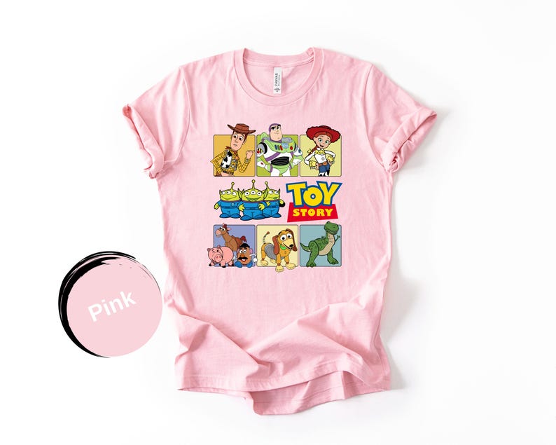 May include: A light pink short-sleeve t-shirt featuring a graphic print of Toy Story characters including Woody, Buzz Lightyear, Jessie, the Aliens, Bullseye, Hamm, Mr. Potato Head, Slinky Dog, and Rex, with the Toy Story logo in the center.