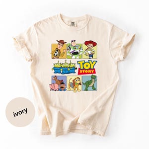 May include: An ivory short-sleeve t-shirt featuring a graphic print of Toy Story characters including Woody, Buzz Lightyear, Jessie, the green aliens, Bullseye, Hamm, Mr. Potato Head, Slinky Dog, and Rex, with the text Toy Story in blue and red.