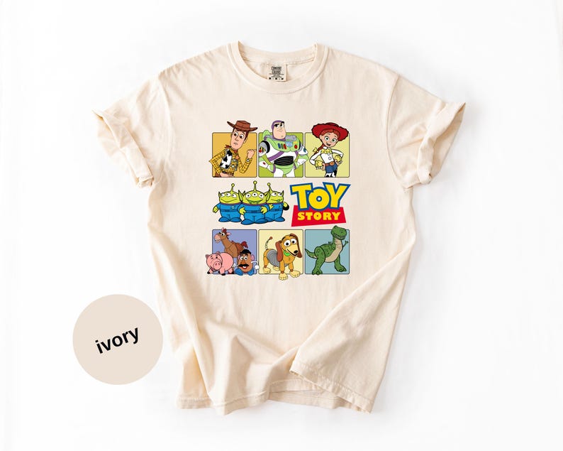 May include: An ivory short-sleeve t-shirt featuring a graphic print of Toy Story characters including Woody, Buzz Lightyear, Jessie, the green aliens, Bullseye, Hamm, Mr. Potato Head, Slinky Dog, and Rex, with the text Toy Story in blue and red.
