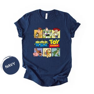 May include: A navy blue short-sleeve t-shirt featuring a graphic print of Toy Story characters including Woody, Buzz Lightyear, Jessie, the Aliens, Bullseye, Hamm, Mr. Potato Head, Slinky Dog, and Rex, with the text Toy Story in yellow and red.