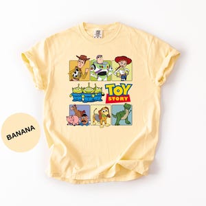 May include: A light yellow t-shirt featuring a graphic print of Toy Story characters including Woody, Buzz Lightyear, Jessie, the Aliens, Bullseye, Hamm, Mr. Potato Head, Slinky Dog, and Rex, with the Toy Story logo text in the center.