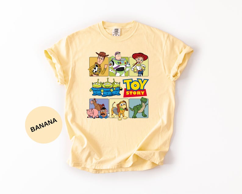 May include: A light yellow t-shirt featuring a graphic print of Toy Story characters including Woody, Buzz Lightyear, Jessie, the Aliens, Bullseye, Hamm, Mr. Potato Head, Slinky Dog, and Rex, with the Toy Story logo text in the center.