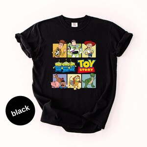 May include: A black short-sleeve t-shirt featuring a graphic print of Toy Story characters including Woody, Buzz Lightyear, Jessie, the Aliens, Bullseye, Hamm, Mr. Potato Head, Slinky Dog, and Rex, with the Toy Story logo in the center.
