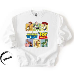May include: A white crewneck sweatshirt featuring a colorful graphic print of Toy Story characters including Woody, Buzz Lightyear, Jessie, the Aliens, Hamm, Mr. Potato Head, Slinky Dog, and Rex, with the Toy Story logo printed in the center.