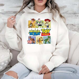 May include: A cream-colored hooded sweatshirt featuring a colorful graphic print of Toy Story characters including Woody, Buzz Lightyear, Jessie, the Aliens, Bullseye, Hamm, Mr. Potato Head, Slinky Dog, and Rex, with the Toy Story logo text.