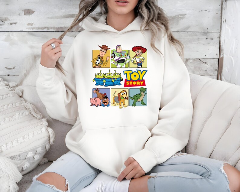 May include: A cream-colored hooded sweatshirt featuring a colorful graphic print of Toy Story characters including Woody, Buzz Lightyear, Jessie, the Aliens, Bullseye, Hamm, Mr. Potato Head, Slinky Dog, and Rex, with the Toy Story logo text.