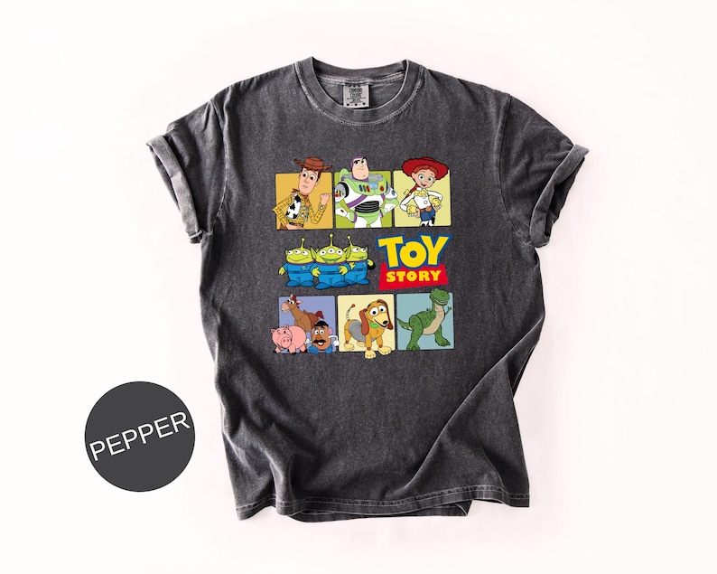 May include: A charcoal gray short-sleeve t-shirt featuring a graphic print of Toy Story characters including Woody, Buzz Lightyear, Jessie, the Aliens, Bullseye, Hamm, Mr. Potato Head, Slinky Dog, and Rex, with the text Toy Story in yellow and red.