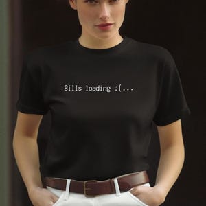 May include: Black t-shirt with the text "Bills loading :(..." in white. The shirt has short sleeves and a crew neck. The model is wearing a brown leather belt and white pants.