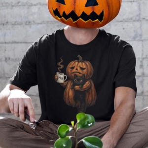 May include: A black t-shirt featuring a graphic of a sad jack-o'-lantern holding a steaming white mug with the text "MEH" printed on it, while also cradling a small black cat. The design is centred on the chest of the short-sleeved garment.