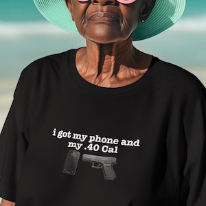 May include: A black short-sleeved t-shirt featuring white text that reads, "i got my phone and my .40 Cal," accompanied by a graphic illustration of a black smartphone and a black handgun positioned directly below the printed text.