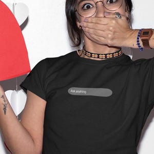May include: A black short-sleeved t-shirt featuring a grey horizontal rectangular graphic on the chest. Inside the grey rectangle, the text "Ask anything" is printed in a small, white, sans-serif font. The shirt has a standard crew neckline.