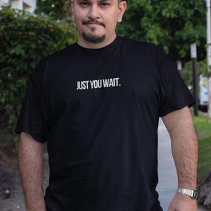 May include: Black t-shirt with the white text "JUST YOU WAIT." The t-shirt is a classic crew neck design. The person wearing the shirt is standing outdoors on a pavement.