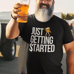 May include: Black t-shirt with the text "JUST GETTING STARTED" in white, featuring a cartoon beer mug. A person is holding a glass of beer. The image is taken outdoors.
