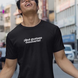 May include: A black short-sleeved t-shirt featuring the white text "¡Qué gustazo conocerte!" printed across the chest. The garment has a classic crew neckline and a standard fit, providing a clean and simple look for casual everyday wear.