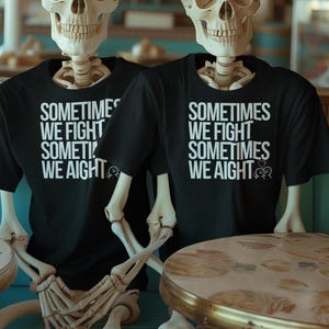 May include: Two black t-shirts with the text "SOMETIMES WE FIGHT SOMETIMES WE AIGHT" in white. The shirts are worn by skeletons sitting at a table. The table has a round top with a gold rim.