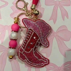 May include: A keychain featuring a cowboy boot and hat shape covered in pink and red rhinestones with silver star accents. It includes a gold-tone metal clasp, a pink tassel, and a strand of alternating pink and white beads with red rhinestone spacers.